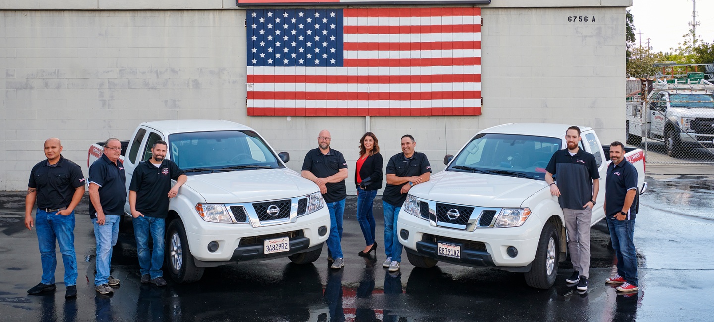 Team of Overhead Door Company of Sacramento employees standing in front of two white Nissan trucks, with an American flag mural in the background, showcasing professionalism and community commitment.