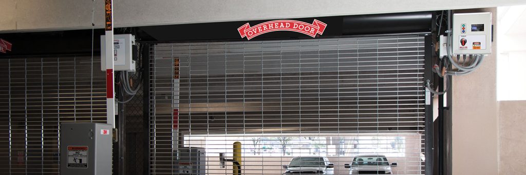 Commercial garage door with Overhead Door branding, featuring a horizontal grille design, control panel, and safety features, set in a parking garage environment.